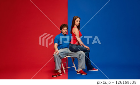 Serious young man and woman, couple sitting together on stool with neutral expressions, against vivid red and blue background. Serious young man and woman, couple sitting together on stool with neutral expressions, against vivid red and blue background. 126169539