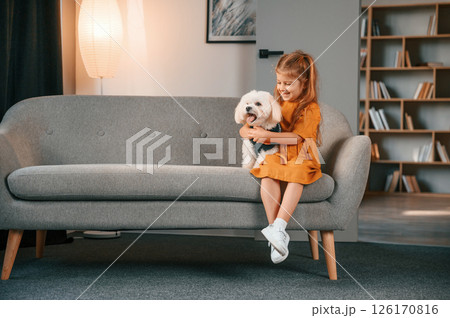 Little girl in yellow clothes is sitting on the sofa with maltese dog indoors in domestic room Little girl in yellow clothes is sitting on the sofa with maltese dog indoors in domestic room 126170816