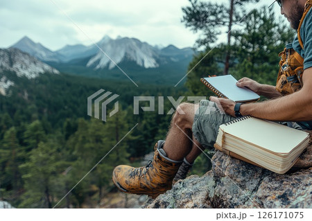 Cropped photo of a man takes notes in a paper notebook while hiking. The man pauses amid breathtaking views, capturing thoughts in his notebook during a memorable hiking experience. 126171075