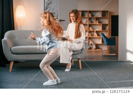 Woman with book, girl is having fun. Mother with her daughter is together in the domestic room 126171124