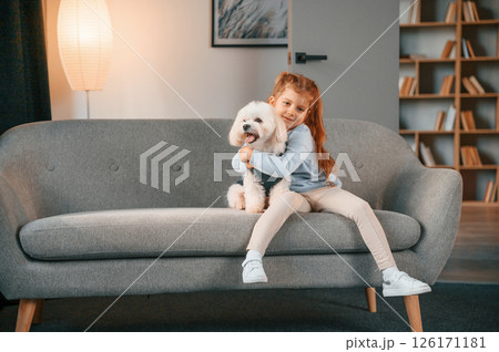Sitting on the sofa. Little girl is with maltese dog indoors in domestic room Sitting on the sofa. Little girl is with maltese dog indoors in domestic room 126171181