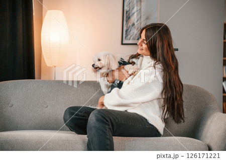 Sitting on the sofa. Beautiful woman is with maltese dog is at home 126171221
