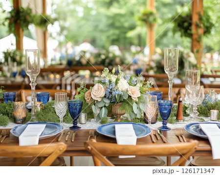 Outdoor banquet in green park with white tablecloth, wooden chairs, floral decor, and elegant settings. 126171347