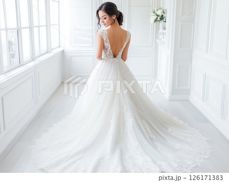 Bride in white dress in bright room, standing back to camera and looking over her shoulder. 126171383