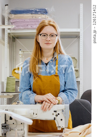 Woman stands in front of a sewing machine, wearing a yellow apron. Woman stands in front of a sewing machine, wearing a yellow apron. 126171412