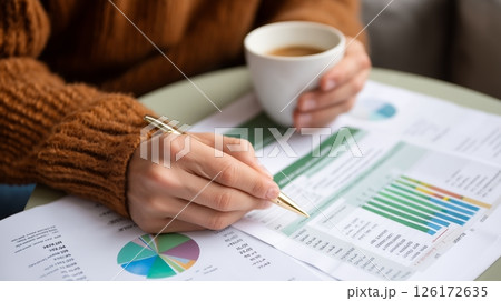 Person analyzes financial reports while sipping coffee in a cozy indoor setting during daytime 126172635