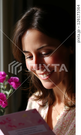Woman enjoying a moment of happiness while reading a card near blooming flowers in a cozy indoor setting during daylight 126173364