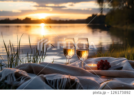 Two glasses of white wine and grapes on a blanket by the lake, creating a romantic atmosphere at sunset Two glasses of white wine and grapes on a blanket by the lake, creating a romantic atmosphere at sunset 126173892