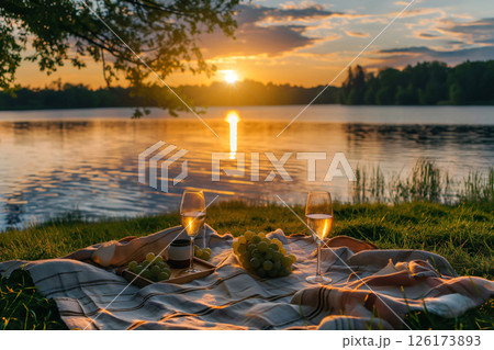 Romantic picnic setting for two by the lake at sunset, featuring a blanket, wine glasses, grapes, and a breathtaking view 126173893