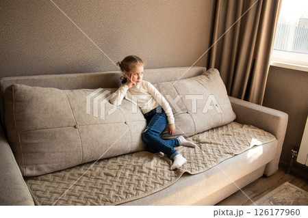 Young girl sitting on cozy sofa wearing casual clothing, with soft textures and warm lighting creating comfortable home atmosphere 126177960