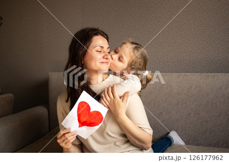 Young girl with light hair hugs mom tightly while holding up her childish drawing. Pure mother-daughter love. 126177962