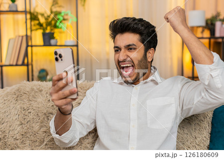 Indian joyful man on sofa happily texting on smartphone, celebrating lottery win, joyful expression 126180609