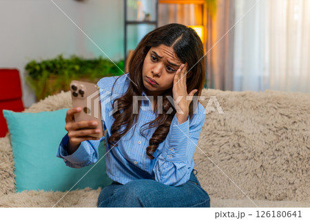 Indian woman looking down with sad sigh while reading bad news on smartphone on home sofa indoors 126180641