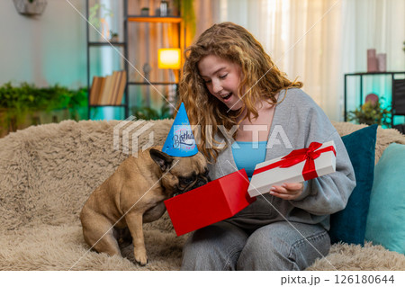 Caucasian woman opens birthday gift on home sofa while pug dog excitedly sniffs box with curiosity Caucasian woman opens birthday gift on home sofa while pug dog excitedly sniffs box with curiosity 126180644