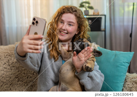 Young woman waving at video call on smartphone with pug dog on home sofa, joyful friendly greeting 126180645