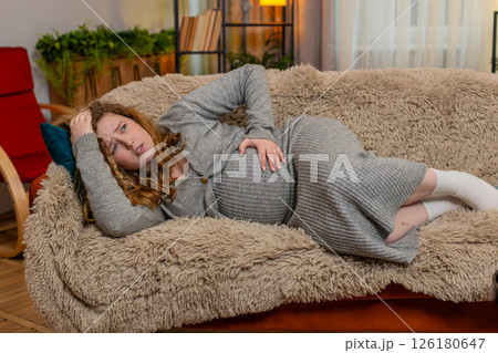 Pregnant young woman lying on home sofa holding belly wincing in pain discomfort worried expression 126180647