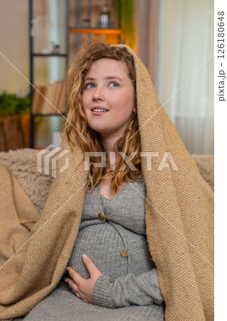 Pregnant young woman wrapped in warm blanket at home gently hugging belly cozy and comforting moment 126180648