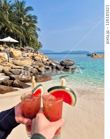 Two hands clinking glasses of watermelon mojito with fruit garnish against stunning Nha Trang beach backdrop. Turquoise sea, white sand, sunny Vietnam paradise Two hands clinking glasses of watermelon mojito with fruit garnish against stunning Nha Trang beach backdrop. Turquoise sea, white sand, sunny Vietnam paradise 126185821