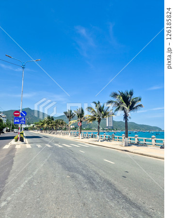 Empty clean road in Nha Trang during siesta time with traffic signs, blue sea featuring traditional Vietnamese boats, palm trees and sunny sky 126185824