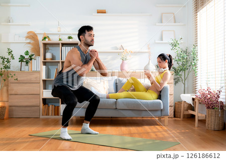 Young asian man doing squat exercise on yoga mat in living room at home while woman using phone. 126186612