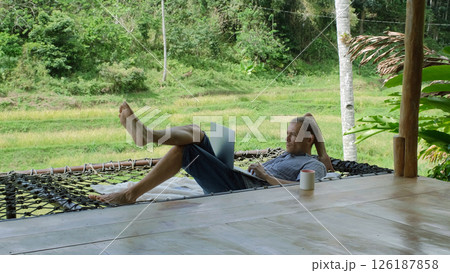 A young man working in a hammock, engaging in remote work on his laptop, surrounded by lush tropical jungle, he enjoys the serenity and beauty of nature on a terrace  126187858