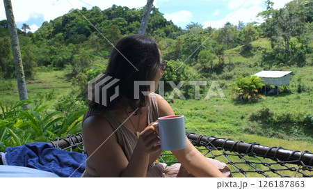 Young woman relaxes in a hammock on a tropical terrace, savoring freshly brewed coffee while taking in breathtaking views of rice fields and lush jungle greenery Young woman relaxes in a hammock on a tropical terrace, savoring freshly brewed coffee while taking in breathtaking views of rice fields and lush jungle greenery 126187863