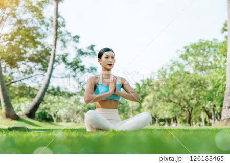Asian woman meditating in a peaceful park setting, sitting cross-legged on green grass with trees 126188465