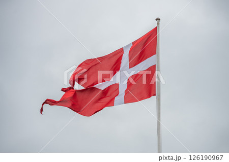 The Denmark flag flutters proudly on a flagpole, displaying its red background and white cross against a cloudy, overcast sky. The atmosphere is calm and reflective. The Denmark flag flutters proudly on a flagpole, displaying its red background and white cross against a cloudy, overcast sky. The atmosphere is calm and reflective. 126190967