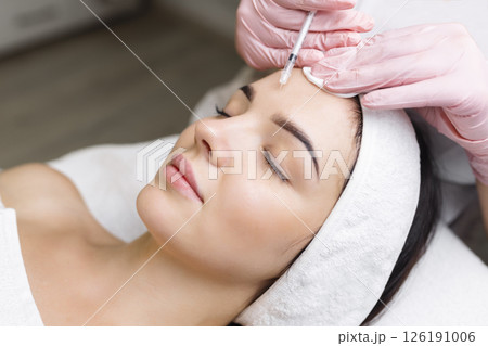 Close up of hands of young cosmetologist injecting botox in female face. She is standing and smiling. The woman is closed her eyes with relaxation 126191006