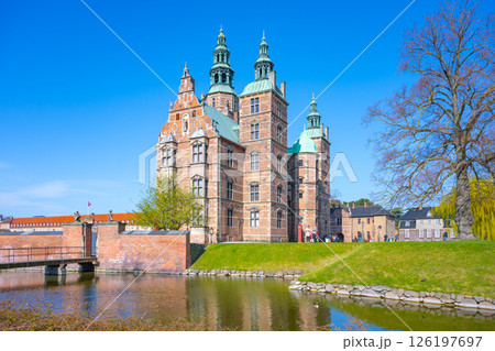 Rosenborg Castle stands majestically by the lake, showcasing its intricate architecture against a clear blue sky. Visitors stroll around the lush gardens enjoying the serene atmosphere. 126197697