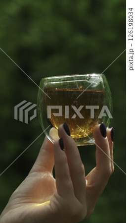 A Hand Holding a Glass Filled with Golden Tea While Surrounded by Beautiful Nature. Media 126198034