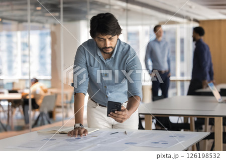 Businessman reviewing information on smartphone standing at desk in office Businessman reviewing information on smartphone standing at desk in office 126198532