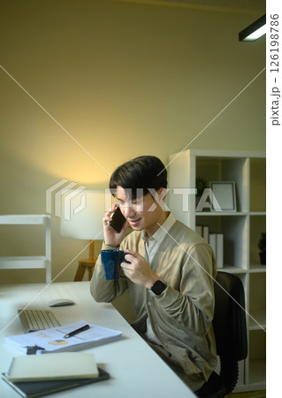 Young entrepreneur holding cup of coffee and talking on mobile phone at home office desk Young entrepreneur holding cup of coffee and talking on mobile phone at home office desk 126198786