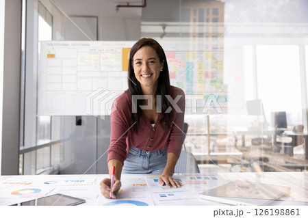 Portrait confident young businesswoman standing by desk filled with papers Portrait confident young businesswoman standing by desk filled with papers 126198861