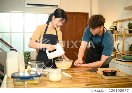 Smiling couple making pancake batter together using a hand mixer in a kitchen Smiling couple making pancake batter together using a hand mixer in a kitchen 126198977