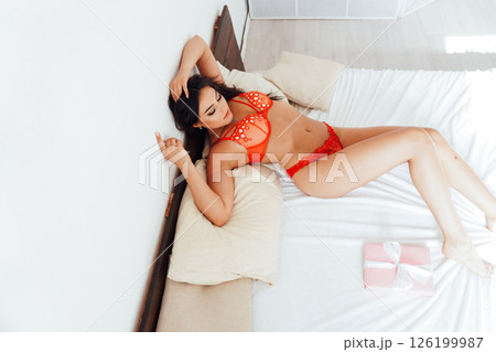 Brunette woman in red lingerie with pink gift in bedroom on bed in morning Brunette woman in red lingerie with pink gift in bedroom on bed in morning 126199987