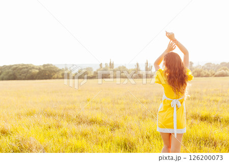 Slender woman in yellow summer dress walking in wheat field at sunset 126200073