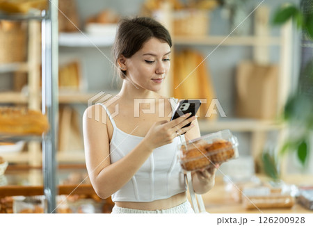 Cute young girl choosing pastries in bakery store, scans QR code on packaging box using mobile 126200928