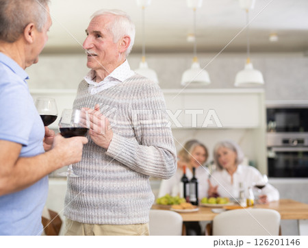 Elderly men have withdrawn from their female companions, stand with glass of wine in hands, chatting 126201146