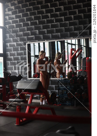 A Woman Using Her Smartphone in the Gym While Looking at Her Reflection in the Mirror 126201844