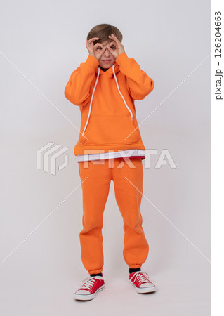 Boy in orange sweatshirt and orange pants making glasses sign with hands. Boy standing on grey background 126204663