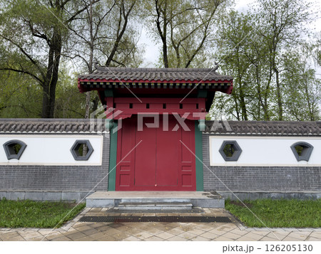 Traditional red wooden gate with tiled roof and geometric windows in decorative wall, inspired by East Asian architecture. Symmetry design and natural park background make. Ideal for visual projects 126205130