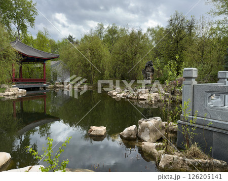 Scenic view of traditional Chinese garden with red pavilion by the pond, stone bridge elements, ornamental rocks and lush greenery reflecting in the still water. Serene and timeless scene Scenic view of traditional Chinese garden with red pavilion by the pond, stone bridge elements, ornamental rocks and lush greenery reflecting in the still water. Serene and timeless scene 126205141