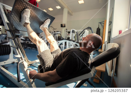 A man is working out in a gym using a leg press machine. He is wearing shorts and a black T-shirt. A man is working out in a gym using a leg press machine. He is wearing shorts and a black T-shirt. 126205387