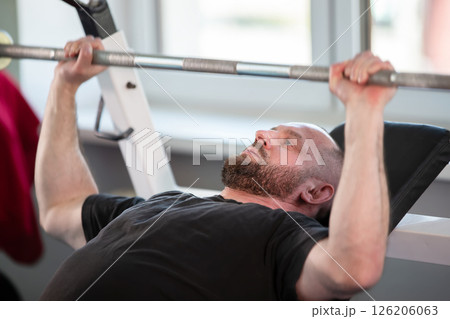 A man is lifting weights on a bench. He is wearing a black shirt and has a beard A man is lifting weights on a bench. He is wearing a black shirt and has a beard 126206063