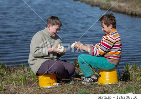 The boys are friends on a fishing trip by the river eating a bun and laughing. Children are friends on a summer fishing trip. 126206510