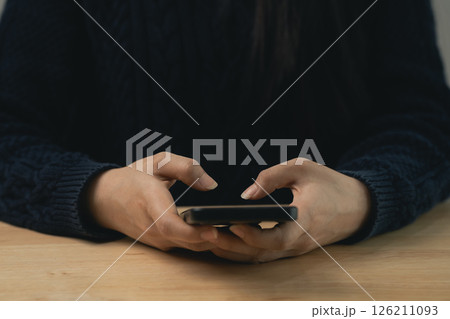 Close-up of Person Holding Smartphone in Hands While Sitting at a Wooden Table with Dark Sweater, Focus on Mobile Device and Fingers 126211093