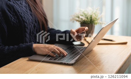 Woman Using Laptop to Make Online Purchase at Home, Holding Credit Card in Hand, Cozy Workspace with Minimalist Decor and Plants 126211094