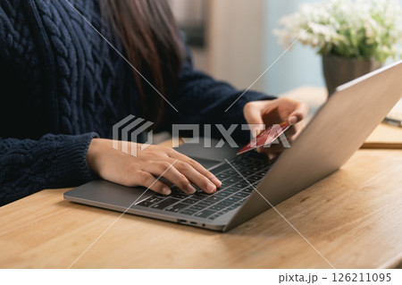 Person using laptop with a credit card for online shopping, sitting at a wooden table, casual indoor environment with decorative plant in background Person using laptop with a credit card for online shopping, sitting at a wooden table, casual indoor environment with decorative plant in background 126211095