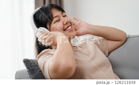Asian woman listening to music happily, having fun and relaxing with headphones in living room. 126211838